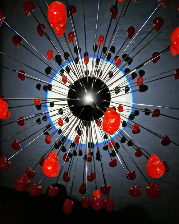 Large spherical hanging sculpture composed of a central black metallic orb with over 70 radiating stainless steel rods, each tipped with a glossy red sculpted skull. The skulls appear to orbit the sphere like a molecular explosion or stylised dandelion, creating a dramatic and surreal visual impact. Materials include stainless steel, polymorph and resin, with vibrant red gloss details and industrial metallic finishes.