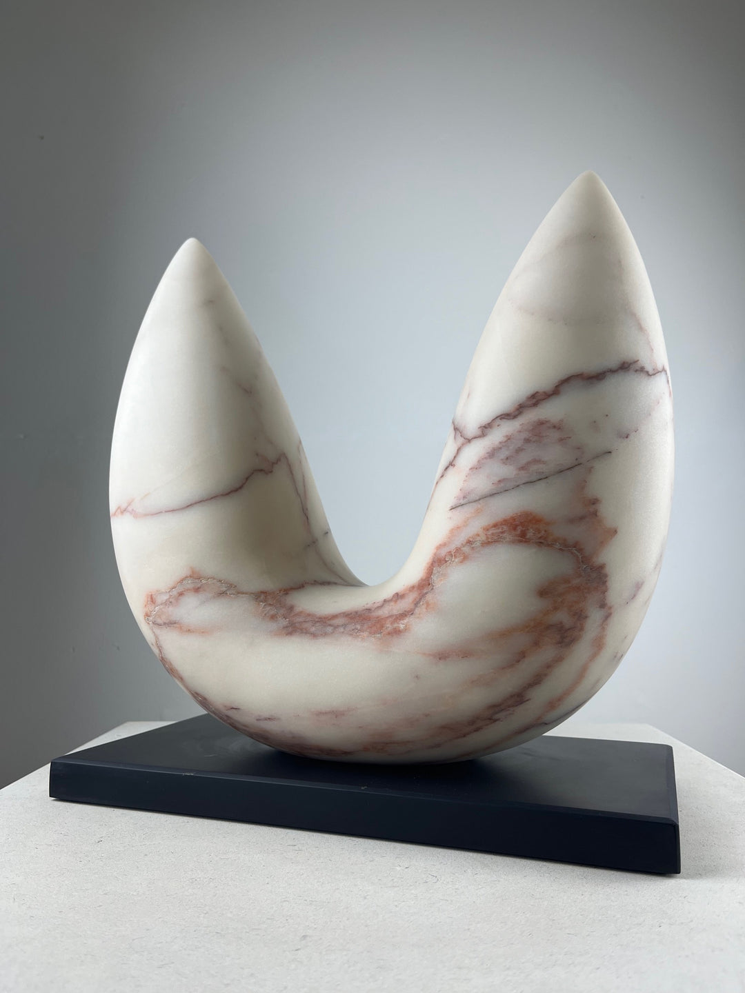 Jason Mulligan sculpture titled FABLE, a one-off abstract form hand-carved from Portuguese marble. The sculpture features a crescent-shaped body rising into two pointed peaks, its smooth surface revealing natural cream tones veined with warm red and soft blush markings. Resting on a dark plinth, the work embodies balance and symbolism, with its upward flow suggesting narrative, myth, and the shared human impulse to shape meaning through form. A contemporary statement piece.