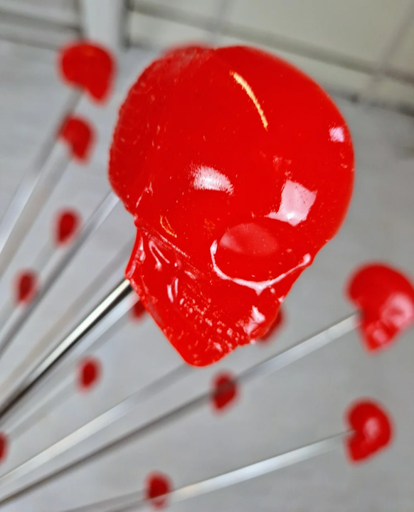 Large spherical hanging sculpture composed of a central black metallic orb with over 70 radiating stainless steel rods, each tipped with a glossy red sculpted skull. The skulls appear to orbit the sphere like a molecular explosion or stylised dandelion, creating a dramatic and surreal visual impact. Materials include stainless steel, polymorph and resin, with vibrant red gloss details and industrial metallic finishes.