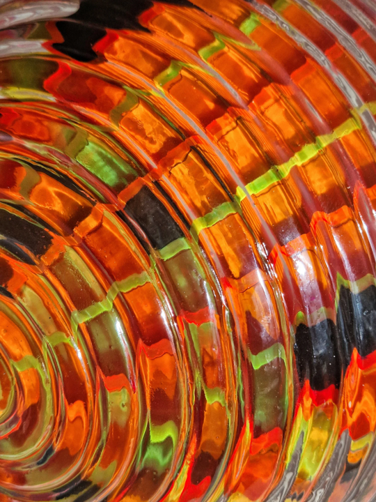 Spherical resin and acrylic sculpture with concentric ridges and vibrant internal colour patterns. The layered composition features fluorescent red, yellow, black and orange acrylic bands encapsulated in clear resin, creating an optical effect that shifts with light and angle. Displayed on a white plinth against a white brick wall, the piece radiates an energetic, cosmic presence—evoking portals, celestial forms, or futuristic artefacts.
