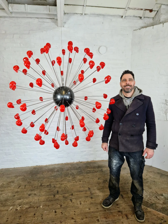 Large spherical hanging sculpture composed of a central black metallic orb with over 70 radiating stainless steel rods, each tipped with a glossy red sculpted skull. The skulls appear to orbit the sphere like a molecular explosion or stylised dandelion, creating a dramatic and surreal visual impact. Materials include stainless steel, polymorph and resin, with vibrant red gloss details and industrial metallic finishes.