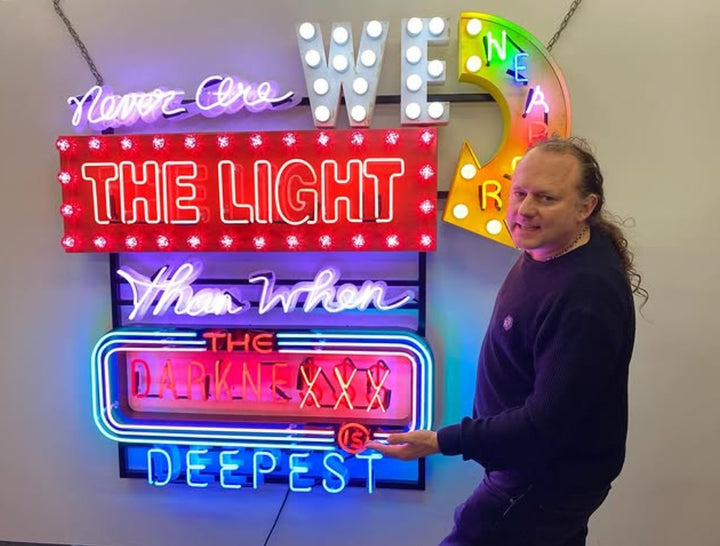 Marcus Bracey with Neon wall artwork featuring the phrase “Never Are We Nearer The Light Than When The Darkness Is Deepest” in layered typography. Includes fabricated metal panels, red marquee lights, a retro arrow lit with cabochon LEDs, and vivid neon lettering in magenta, violet, red, blue, turquoise, orange, and white. A glowing, multicoloured composition by Chris Bracey reflecting spiritual insight and emotional depth.