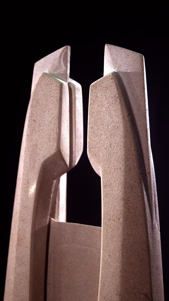 Stone sculpture featuring two tall, curved spires carved from Purbeck Thornback stone, set on a polished black granite base, connected by a narrow central opening with stainless steel elements, displayed against a dark background.