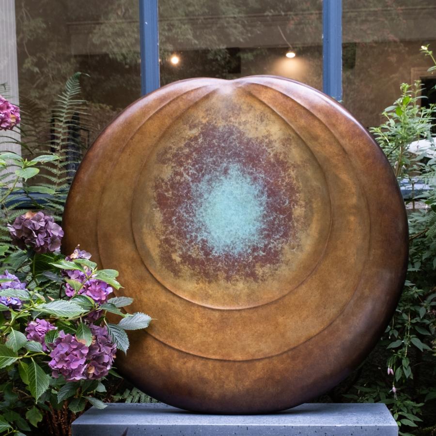 Circular bronze sculpture by Dominic Welch, Aurorean Moon, featuring a layered, concentric form with a glowing turquoise-green patina at its centre, radiating outwards into rich bronze and earthy red-gold tones. A refined limited-edition work blending celestial inspiration with organic presence.