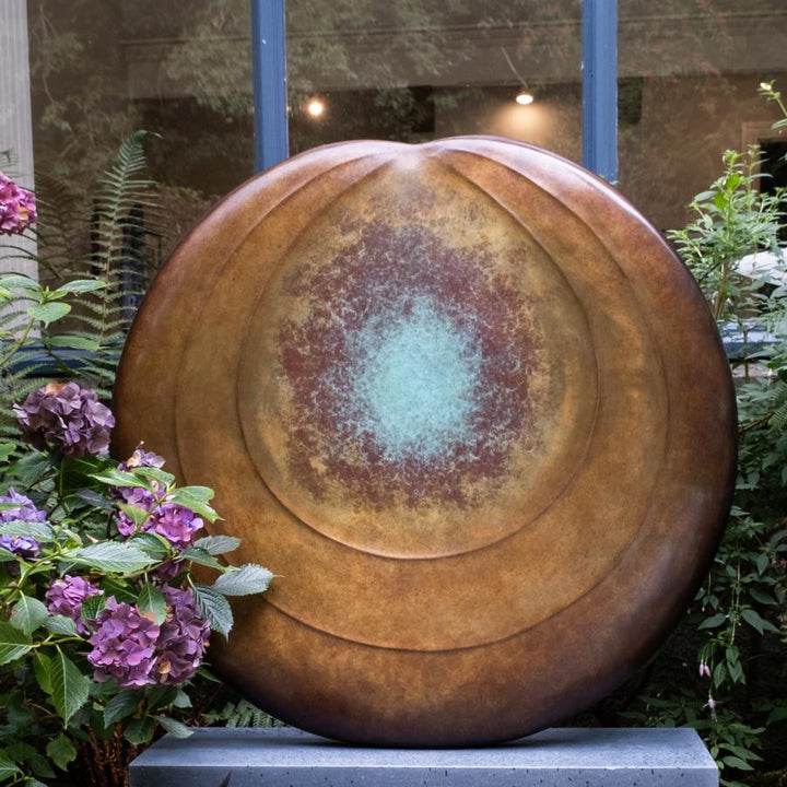 Circular bronze sculpture by Dominic Welch, Aurorean Moon, featuring a layered, concentric form with a glowing turquoise-green patina at its centre, radiating outwards into rich bronze and earthy red-gold tones. A refined limited-edition work blending celestial inspiration with organic presence.
