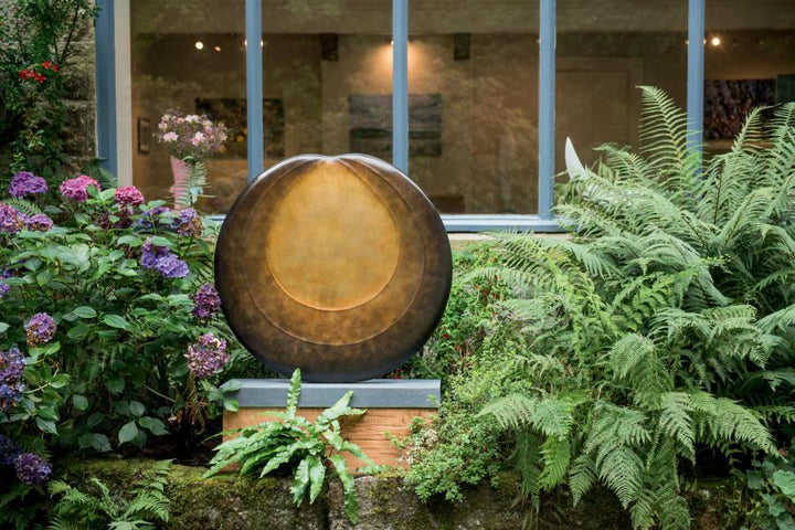 Circular bronze sculpture by Dominic Welch, Aurorean Moon, featuring a layered, concentric form with a glowing turquoise-green patina at its centre, radiating outwards into rich bronze and earthy red-gold tones. A refined limited-edition work blending celestial inspiration with organic presence.