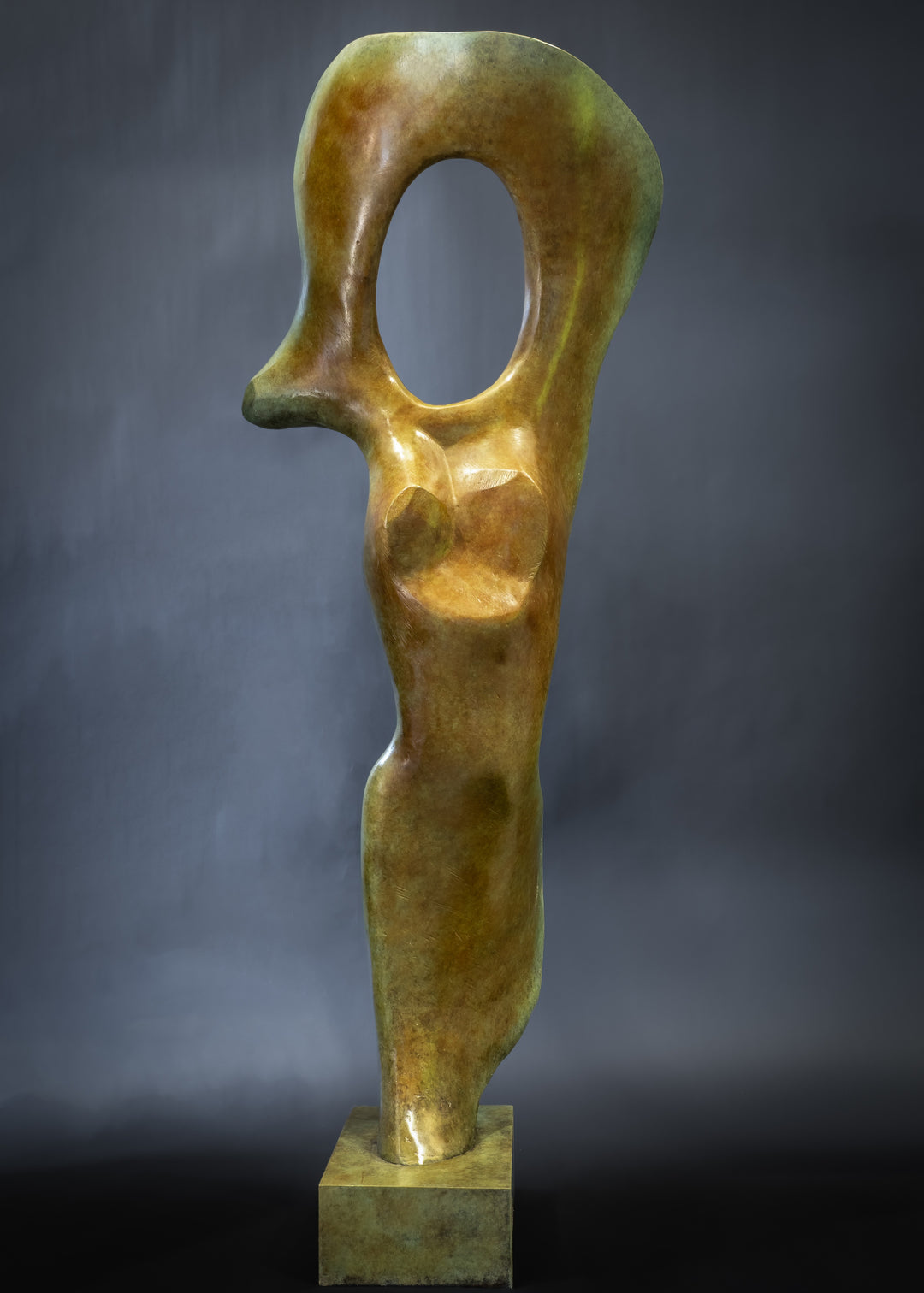 Abstract bronze sculpture of a stylised female torso with flowing curves and a hollowed circular form near the head, displayed in various outdoor and indoor settings. The sculpture has a rich patinated surface with green, gold, and bronze tones.