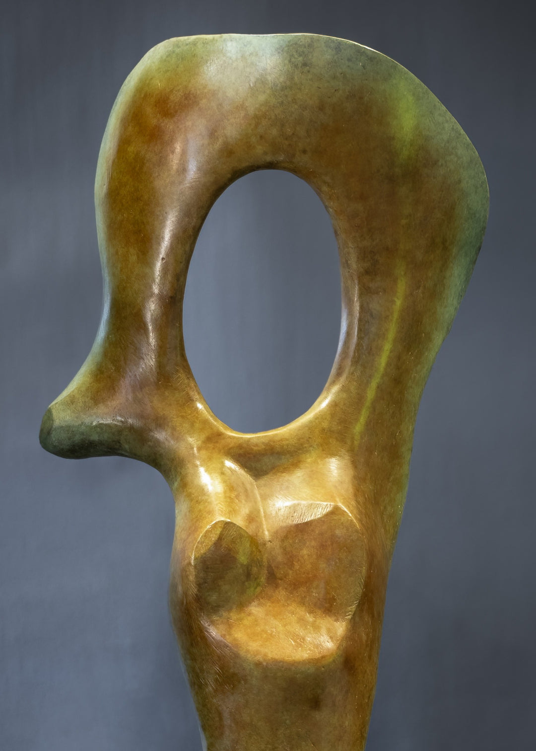 Abstract bronze sculpture of a stylised female torso with flowing curves and a hollowed circular form near the head, displayed in various outdoor and indoor settings. The sculpture has a rich patinated surface with green, gold, and bronze tones.