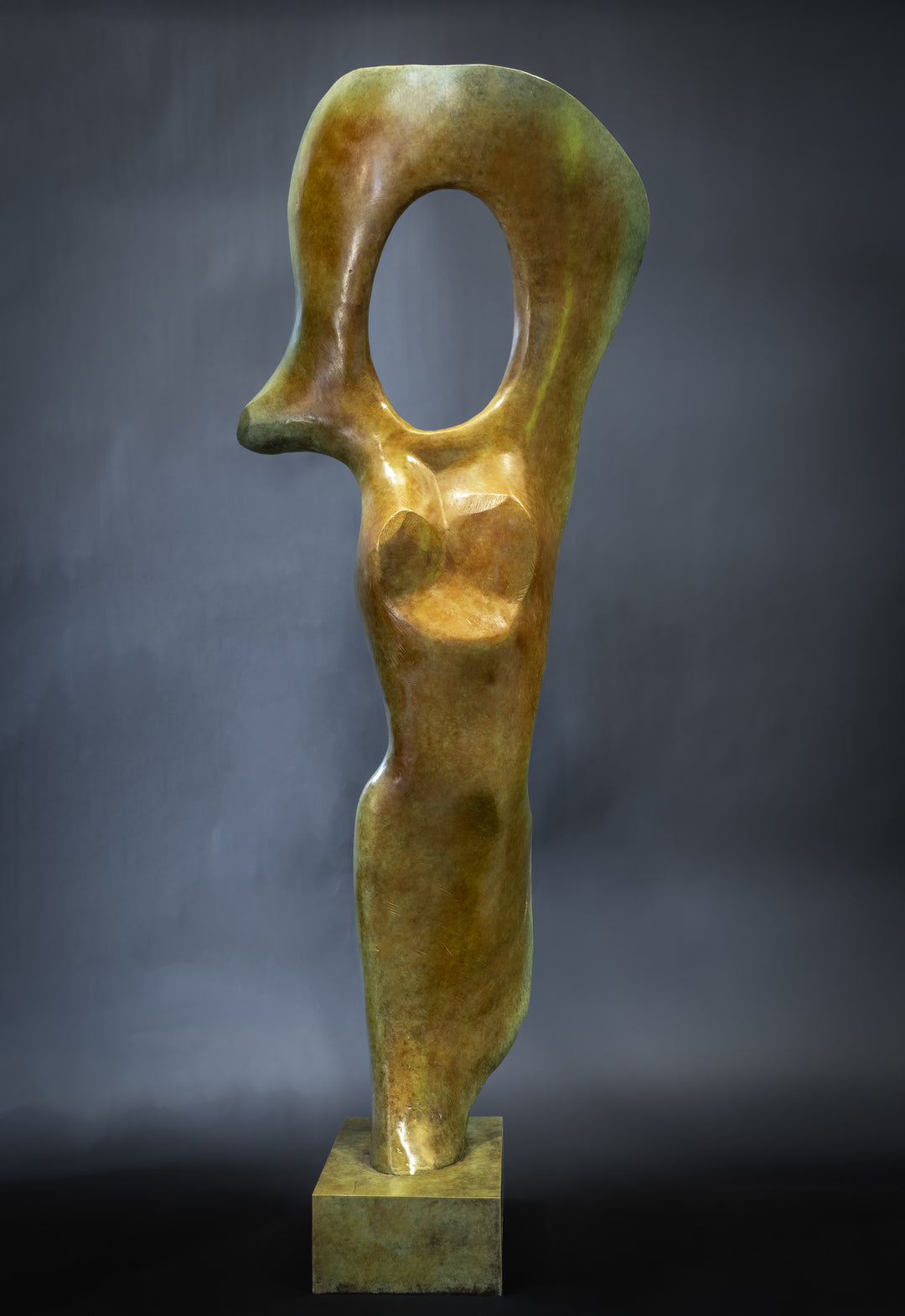 Abstract bronze sculpture of a stylised female torso with flowing curves and a hollowed circular form near the head, displayed in various outdoor and indoor settings. The sculpture has a rich patinated surface with green, gold, and bronze tones.