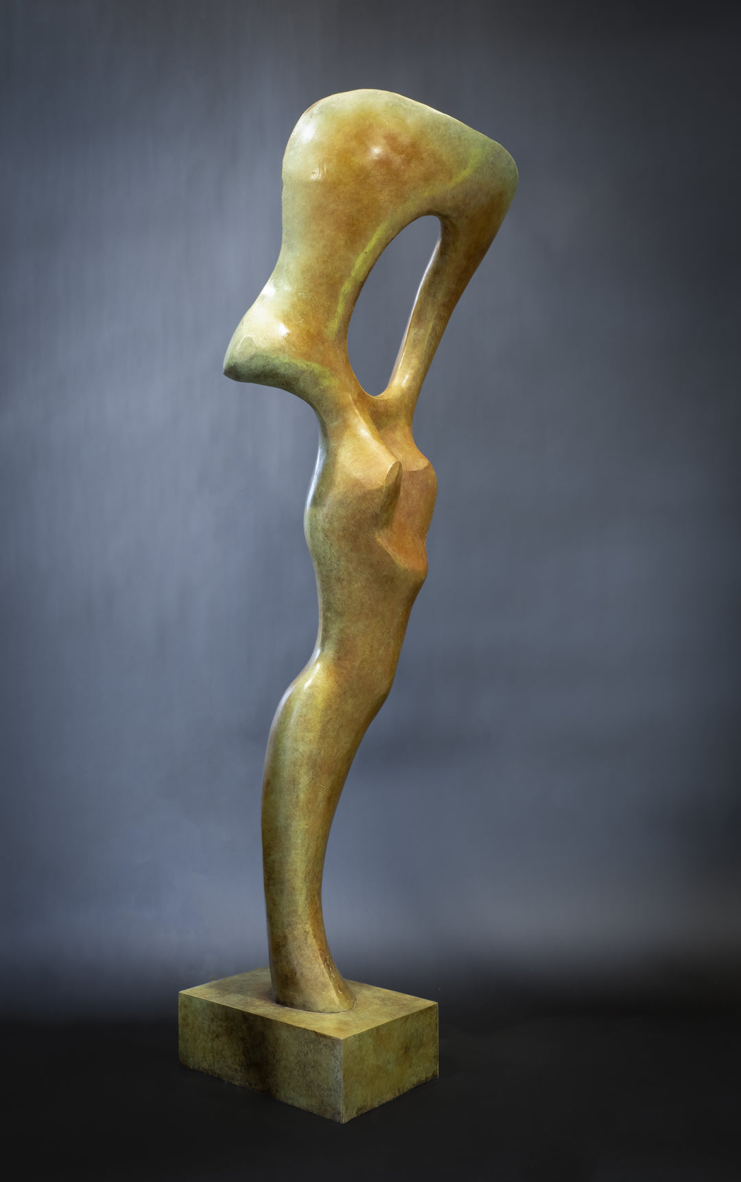 Abstract bronze sculpture of a stylised female torso with flowing curves and a hollowed circular form near the head, displayed in various outdoor and indoor settings. The sculpture has a rich patinated surface with green, gold, and bronze tones.