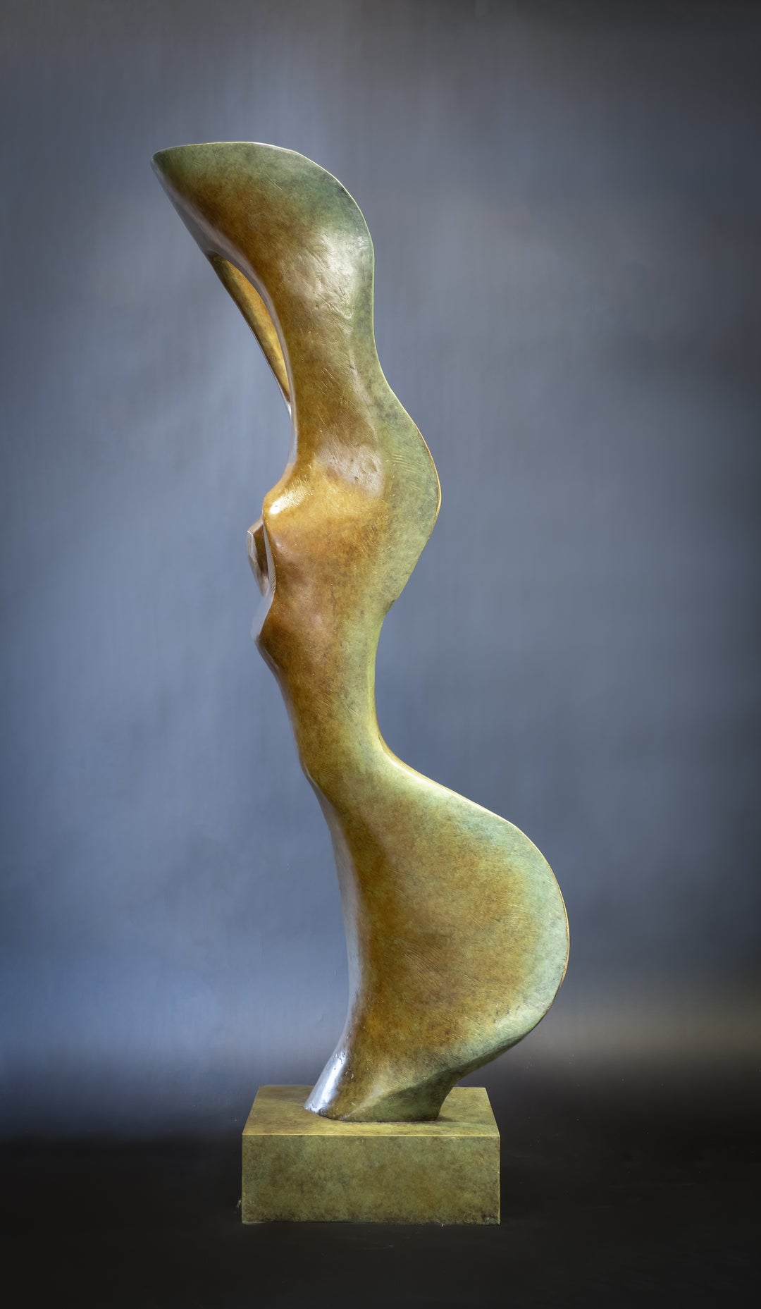 Abstract bronze sculpture of a stylised female torso with flowing curves and a hollowed circular form near the head, displayed in various outdoor and indoor settings. The sculpture has a rich patinated surface with green, gold, and bronze tones.