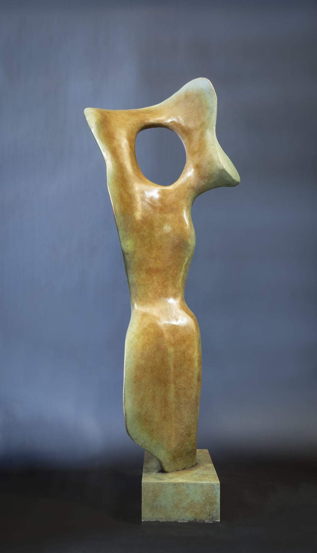 Abstract bronze sculpture of a stylised female torso with flowing curves and a hollowed circular form near the head, displayed in various outdoor and indoor settings. The sculpture has a rich patinated surface with green, gold, and bronze tones.