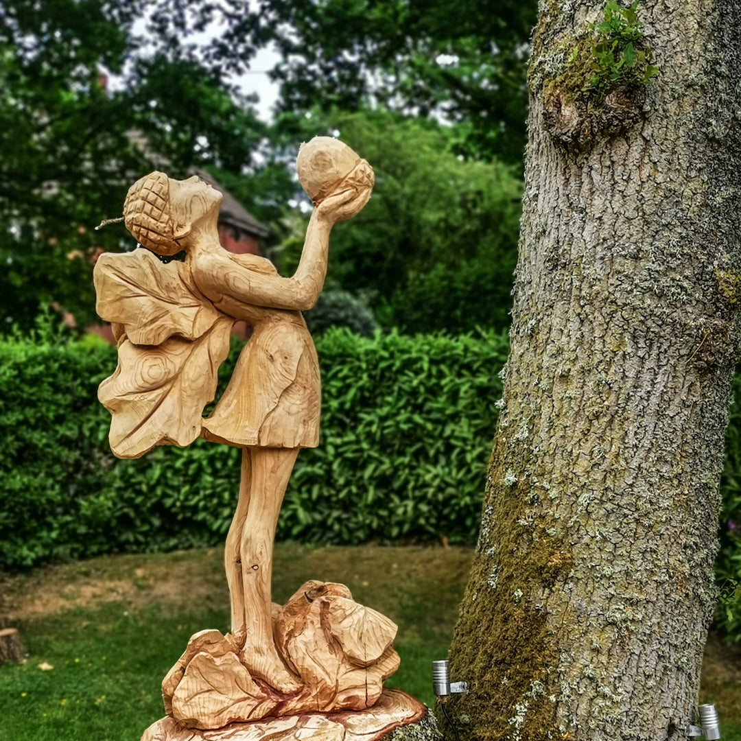 Whimsical wooden sculpture of a fairy standing on a base of carved rocks, holding an acorn aloft and wearing a dress with leaf-shaped wings and an acorn cap, set in a lush garden environment.