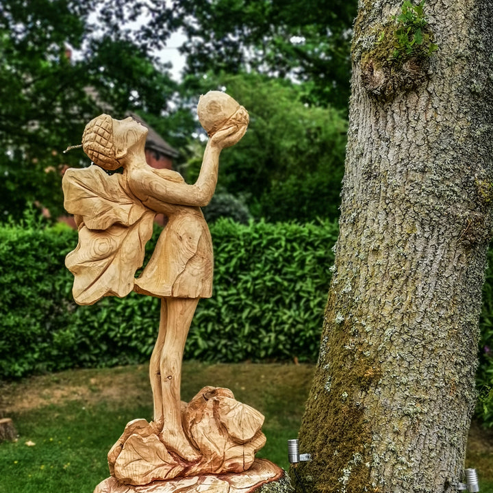 Whimsical wooden sculpture of a fairy standing on a base of carved rocks, holding an acorn aloft and wearing a dress with leaf-shaped wings and an acorn cap, set in a lush garden environment.