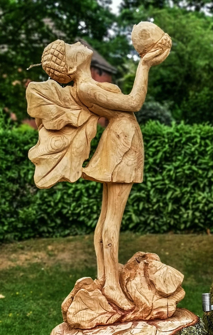 Whimsical wooden sculpture of a fairy standing on a base of carved rocks, holding an acorn aloft and wearing a dress with leaf-shaped wings and an acorn cap, set in a lush garden environment.