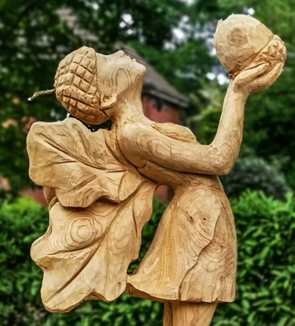 Whimsical wooden sculpture of a fairy standing on a base of carved rocks, holding an acorn aloft and wearing a dress with leaf-shaped wings and an acorn cap, set in a lush garden environment.
