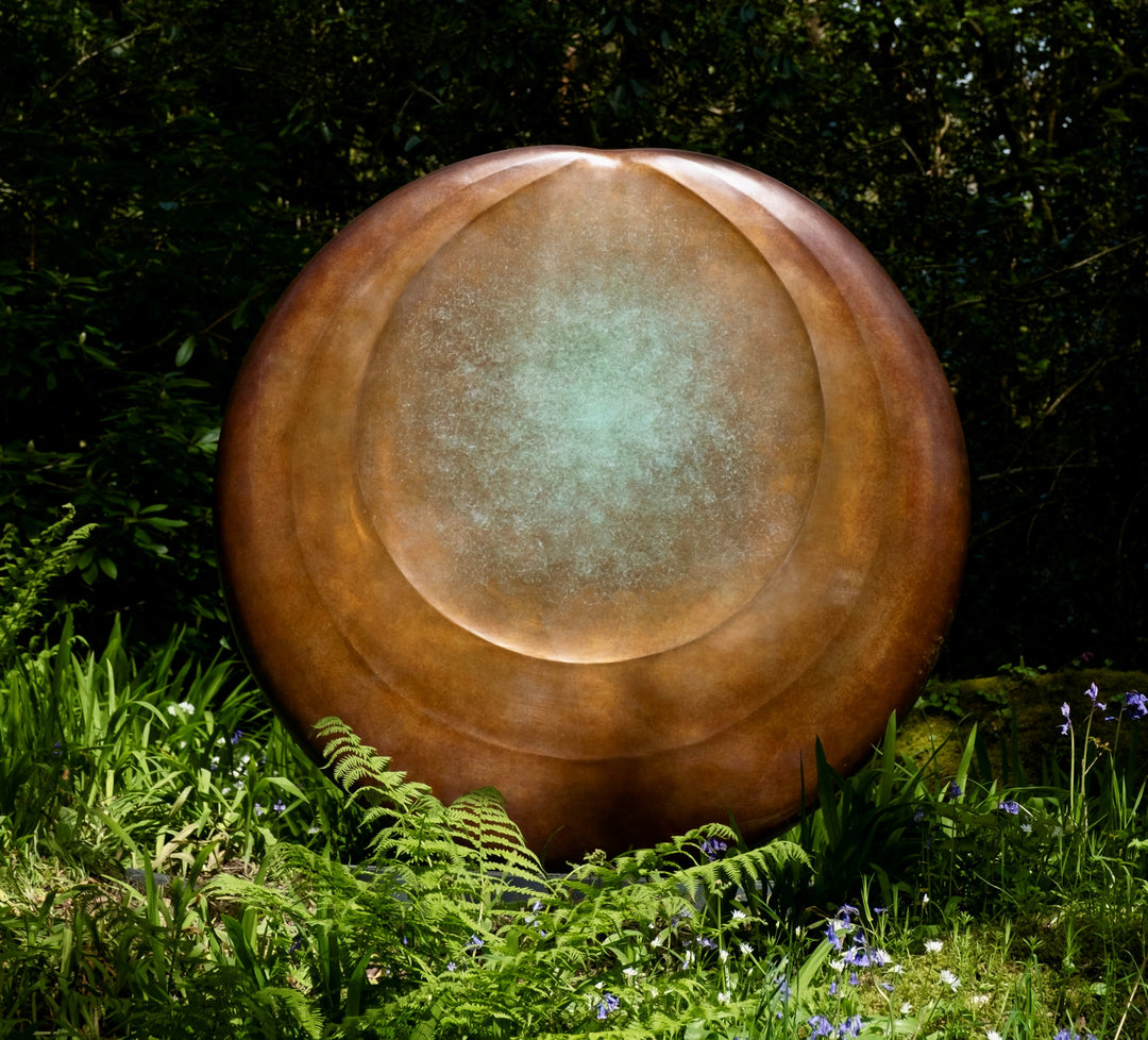 Large circular bronze sculpture Aegean Moon by Dominic Welch, with a luminous verdigris centre fading into warm bronze tones. The surface has subtle textural rings, evoking the phases of the moon. Displayed outdoors among woodland and seasonal landscapes, the piece radiates presence and calm.