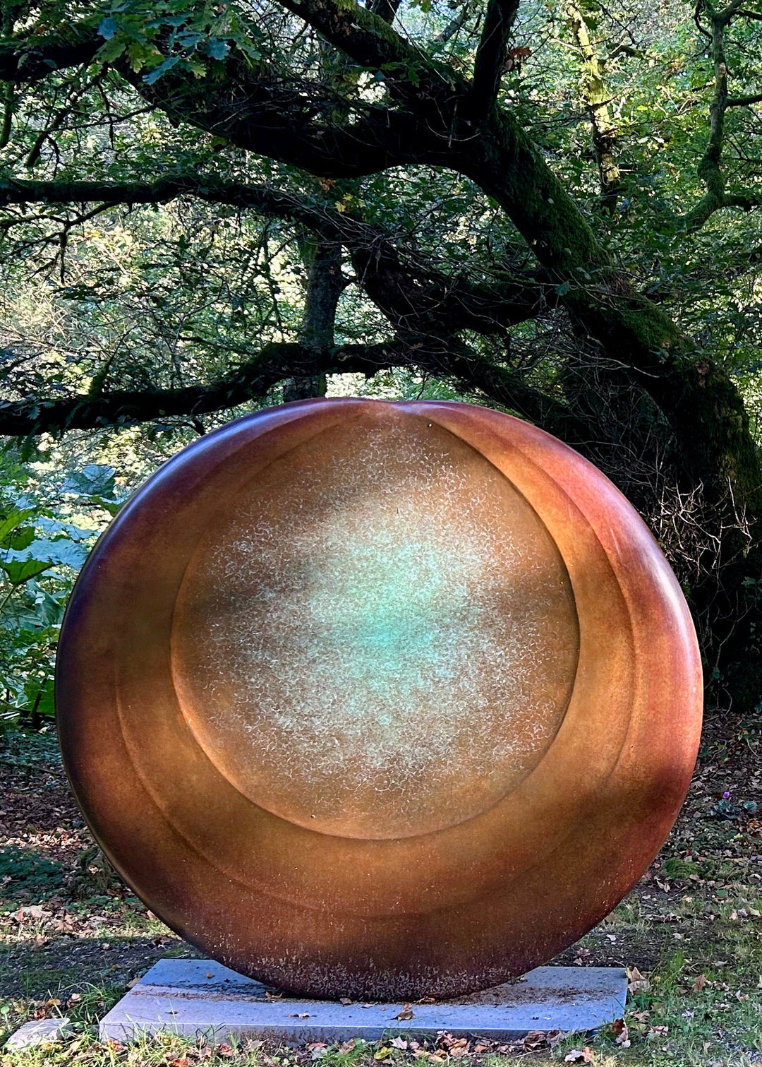 Large circular bronze sculpture Aegean Moon by Dominic Welch, with a luminous verdigris centre fading into warm bronze tones. The surface has subtle textural rings, evoking the phases of the moon. Displayed outdoors among woodland and seasonal landscapes, the piece radiates presence and calm.