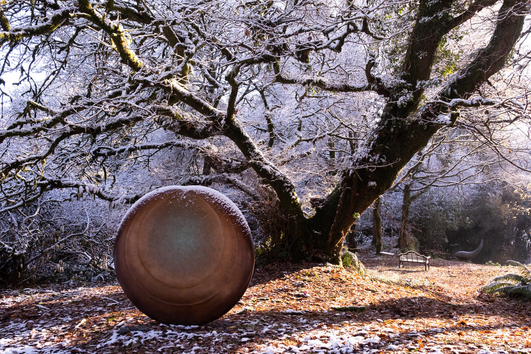 Large circular bronze sculpture Aegean Moon by Dominic Welch, with a luminous verdigris centre fading into warm bronze tones. The surface has subtle textural rings, evoking the phases of the moon. Displayed outdoors among woodland and seasonal landscapes, the piece radiates presence and calm.