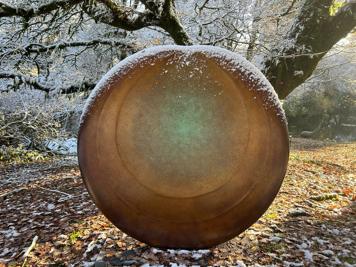 Large circular bronze sculpture Aegean Moon by Dominic Welch, with a luminous verdigris centre fading into warm bronze tones. The surface has subtle textural rings, evoking the phases of the moon. Displayed outdoors among woodland and seasonal landscapes, the piece radiates presence and calm.