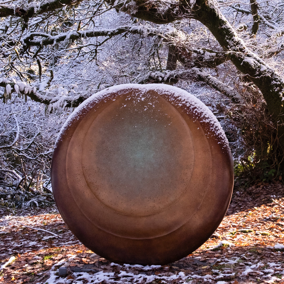 Large circular bronze sculpture Aegean Moon by Dominic Welch, with a luminous verdigris centre fading into warm bronze tones. The surface has subtle textural rings, evoking the phases of the moon. Displayed outdoors among woodland and seasonal landscapes, the piece radiates presence and calm.