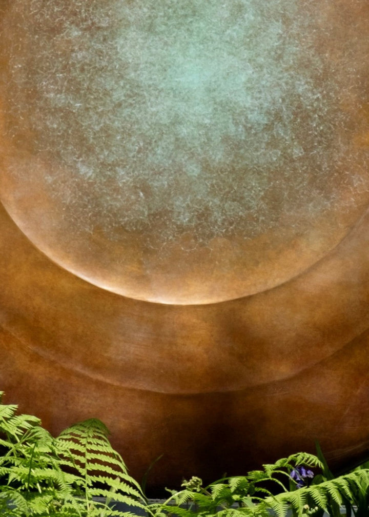 Large circular bronze sculpture Aegean Moon by Dominic Welch, with a luminous verdigris centre fading into warm bronze tones. The surface has subtle textural rings, evoking the phases of the moon. Displayed outdoors among woodland and seasonal landscapes, the piece radiates presence and calm.