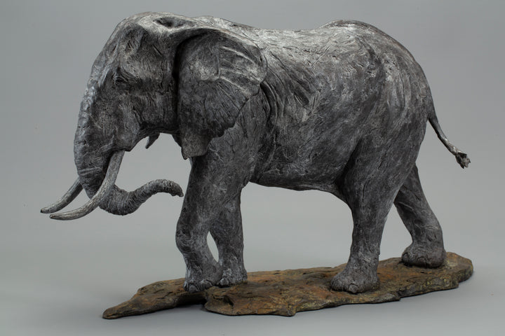 Sculpted elephant in textured grey bronze, shown mid-step with detailed tusks and wrinkled surface, mounted on a naturalistic base
