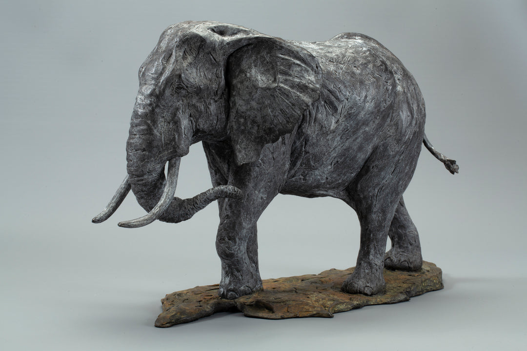 Sculpted elephant in textured grey bronze, shown mid-step with detailed tusks and wrinkled surface, mounted on a naturalistic base