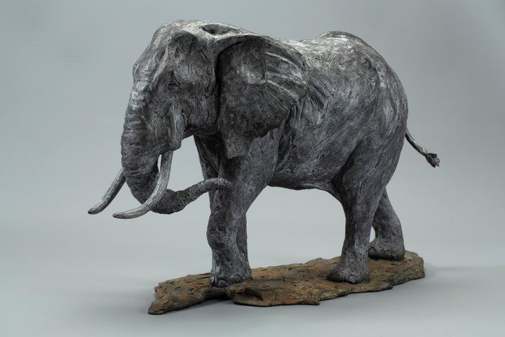 Sculpted elephant in textured grey bronze, shown mid-step with detailed tusks and wrinkled surface, mounted on a naturalistic base