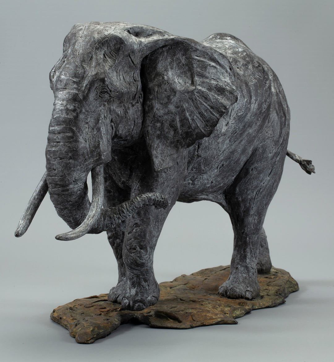 Sculpted elephant in textured grey bronze, shown mid-step with detailed tusks and wrinkled surface, mounted on a naturalistic base