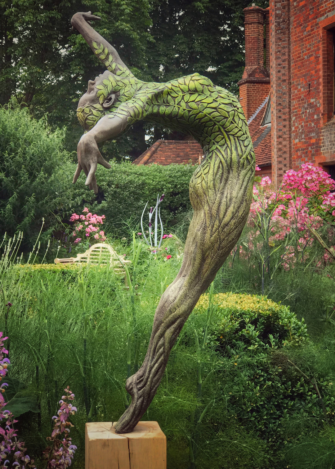 Bronze sculpture of a slender female figure arched forward in a powerful, elongated pose, balanced on one foot atop a wooden plinth. Her body is patterned with leaf-like, cellular shapes that flow from head to toe, echoing natural growth and organic energy. Photographed outdoors among lush green ferns and tall woodland trees.