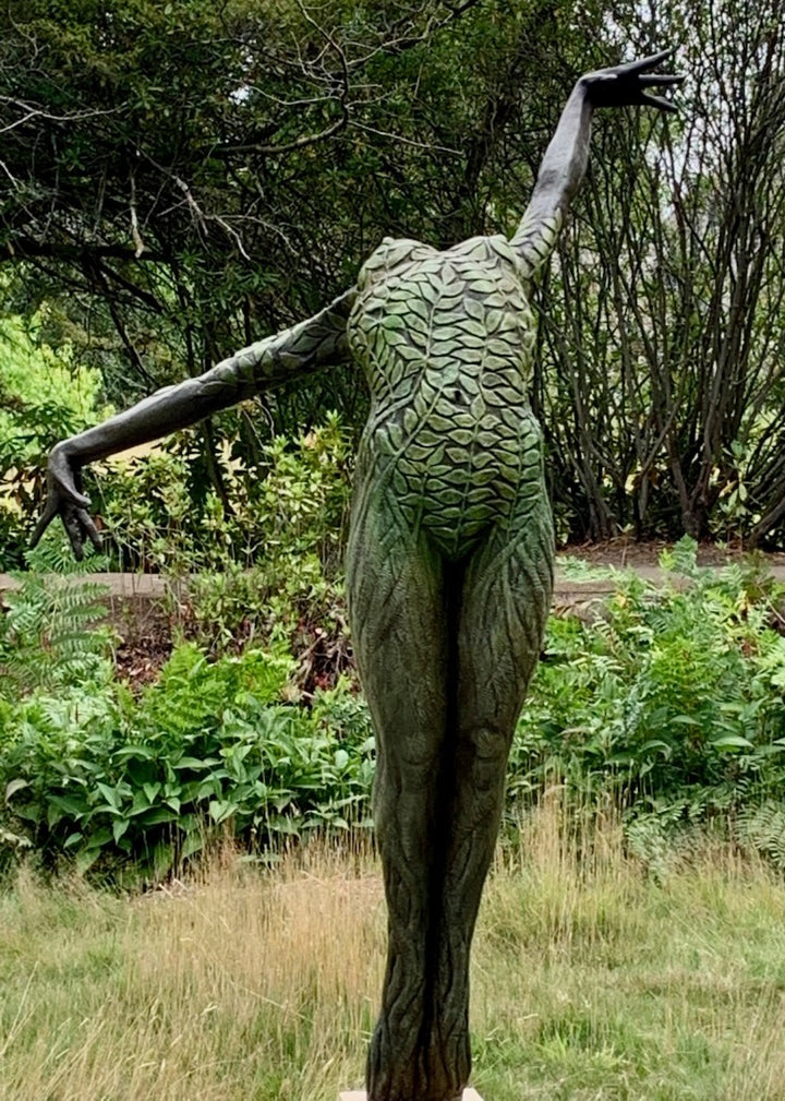Bronze sculpture of a slender female figure arched forward in a powerful, elongated pose, balanced on one foot atop a wooden plinth. Her body is patterned with leaf-like, cellular shapes that flow from head to toe, echoing natural growth and organic energy. Photographed outdoors among lush green ferns and tall woodland trees.