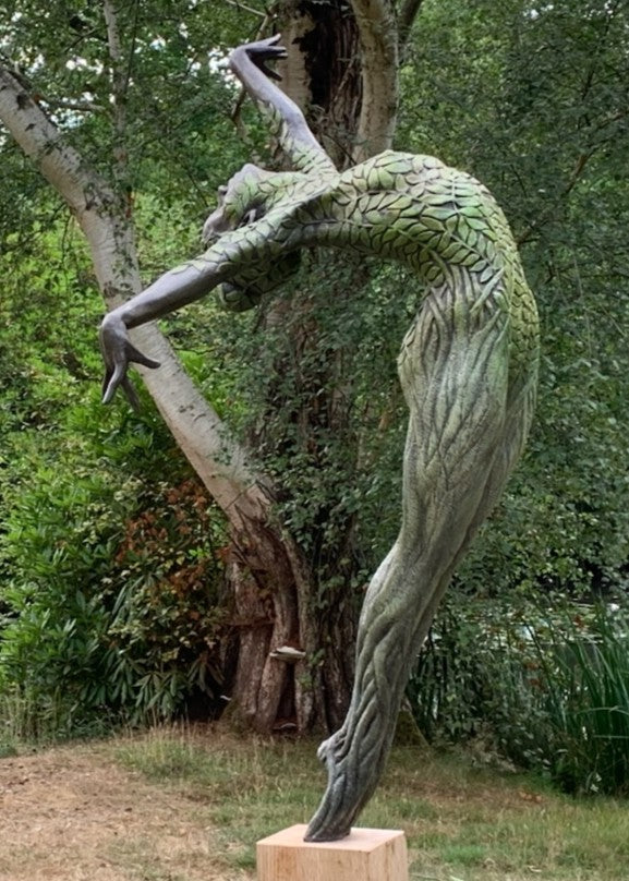 Bronze sculpture of a slender female figure arched forward in a powerful, elongated pose, balanced on one foot atop a wooden plinth. Her body is patterned with leaf-like, cellular shapes that flow from head to toe, echoing natural growth and organic energy. Photographed outdoors among lush green ferns and tall woodland trees.