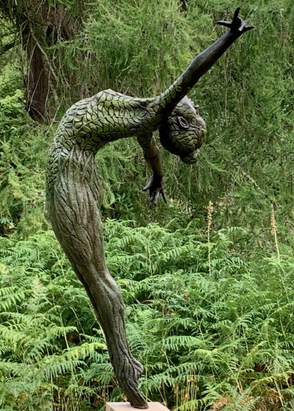 Bronze sculpture of a slender female figure arched forward in a powerful, elongated pose, balanced on one foot atop a wooden plinth. Her body is patterned with leaf-like, cellular shapes that flow from head to toe, echoing natural growth and organic energy. Photographed outdoors among lush green ferns and tall woodland trees.