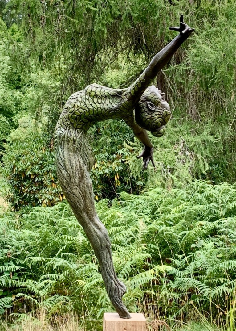 Bronze sculpture of a slender female figure arched forward in a powerful, elongated pose, balanced on one foot atop a wooden plinth. Her body is patterned with leaf-like, cellular shapes that flow from head to toe, echoing natural growth and organic energy. Photographed outdoors among lush green ferns and tall woodland trees.