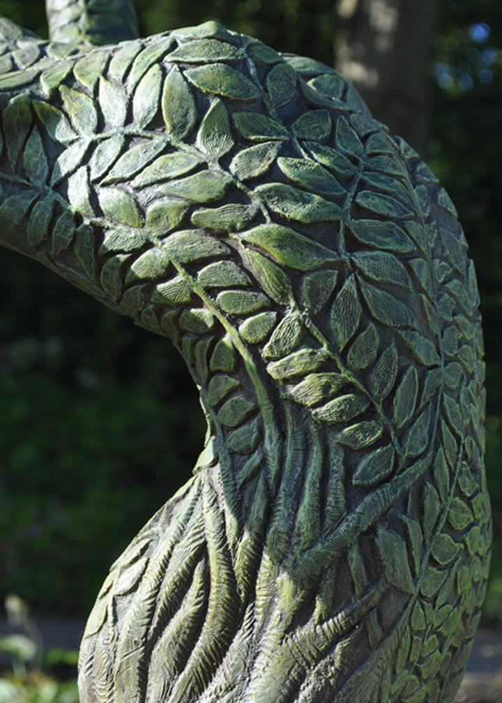 Bronze sculpture of a slender female figure arched forward in a powerful, elongated pose, balanced on one foot atop a wooden plinth. Her body is patterned with leaf-like, cellular shapes that flow from head to toe, echoing natural growth and organic energy. Photographed outdoors among lush green ferns and tall woodland trees.