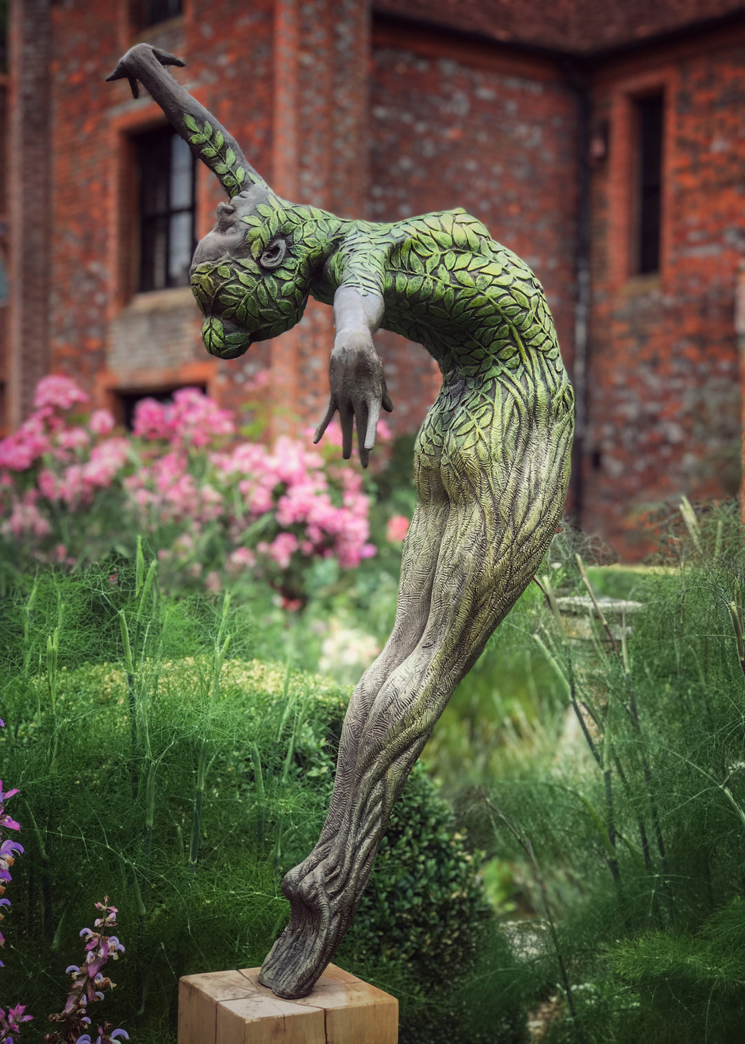 Bronze sculpture of a slender female figure arched forward in a powerful, elongated pose, balanced on one foot atop a wooden plinth. Her body is patterned with leaf-like, cellular shapes that flow from head to toe, echoing natural growth and organic energy. Photographed outdoors among lush green ferns and tall woodland trees.
