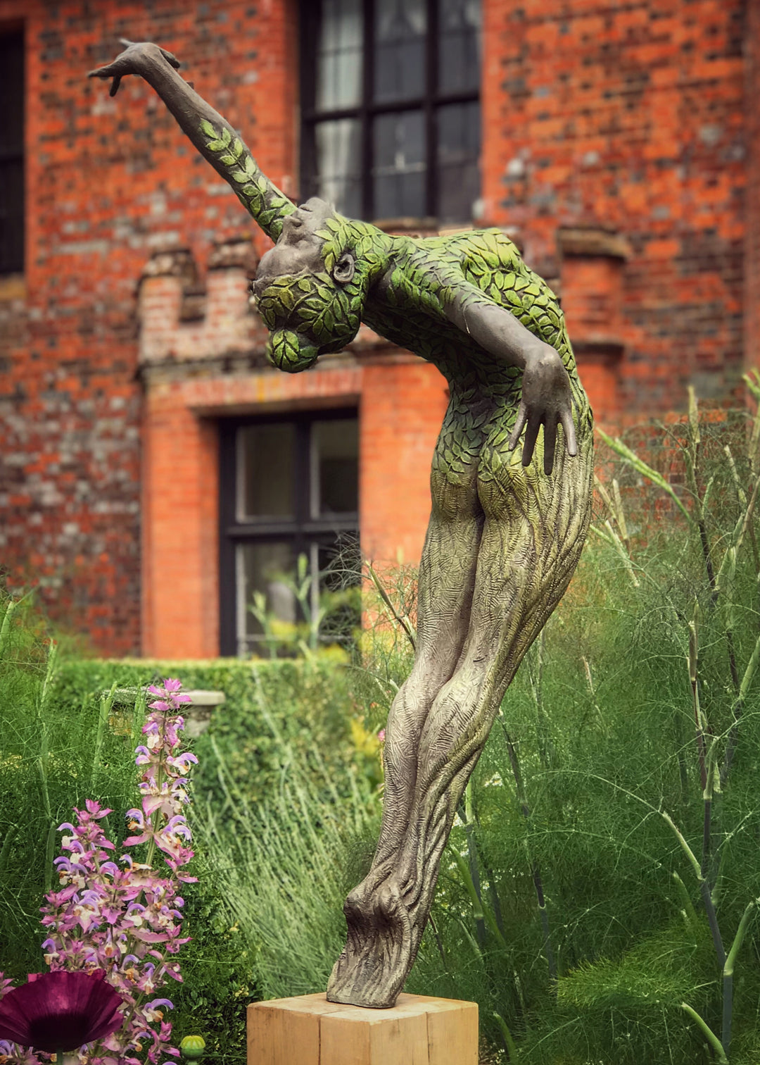 Bronze sculpture of a slender female figure arched forward in a powerful, elongated pose, balanced on one foot atop a wooden plinth. Her body is patterned with leaf-like, cellular shapes that flow from head to toe, echoing natural growth and organic energy. Photographed outdoors among lush green ferns and tall woodland trees.