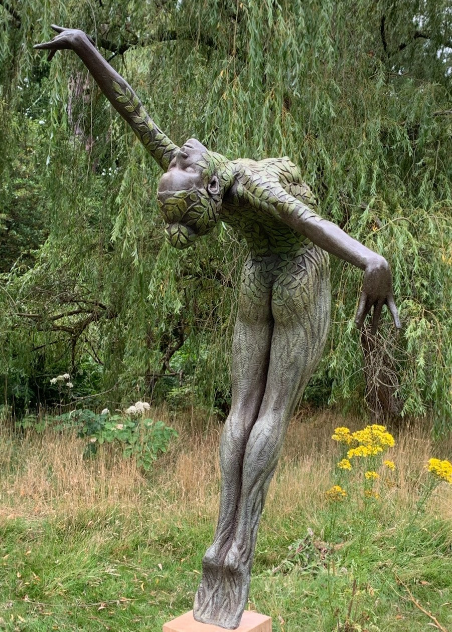 Bronze sculpture of a slender female figure arched forward in a powerful, elongated pose, balanced on one foot atop a wooden plinth. Her body is patterned with leaf-like, cellular shapes that flow from head to toe, echoing natural growth and organic energy. Photographed outdoors among lush green ferns and tall woodland trees.