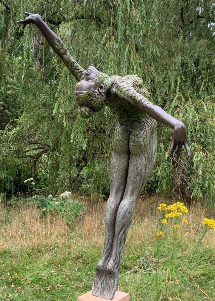 Bronze sculpture of a slender female figure arched forward in a powerful, elongated pose, balanced on one foot atop a wooden plinth. Her body is patterned with leaf-like, cellular shapes that flow from head to toe, echoing natural growth and organic energy. Photographed outdoors among lush green ferns and tall woodland trees.