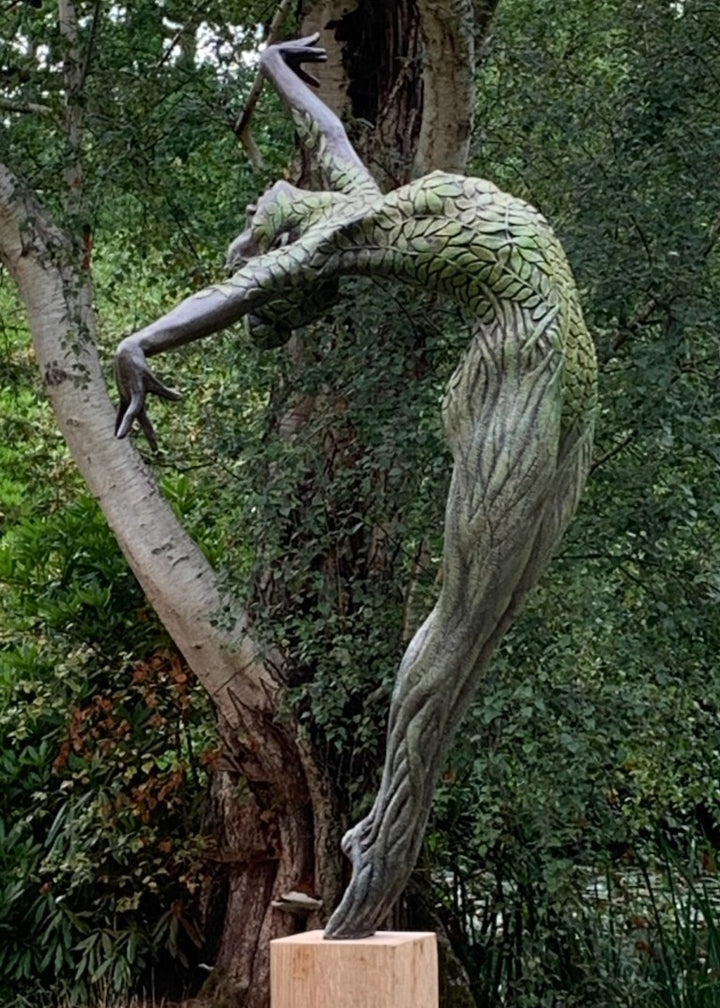 Bronze sculpture of a slender female figure arched forward in a powerful, elongated pose, balanced on one foot atop a wooden plinth. Her body is patterned with leaf-like, cellular shapes that flow from head to toe, echoing natural growth and organic energy. Photographed outdoors among lush green ferns and tall woodland trees.