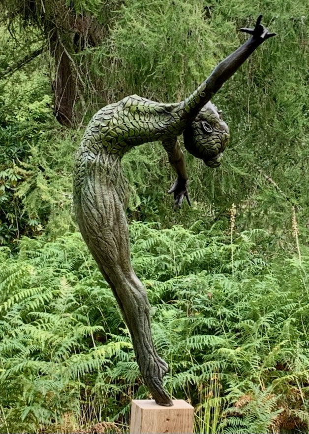 Bronze sculpture of a slender female figure arched forward in a powerful, elongated pose, balanced on one foot atop a wooden plinth. Her body is patterned with leaf-like, cellular shapes that flow from head to toe, echoing natural growth and organic energy. Photographed outdoors among lush green ferns and tall woodland trees.