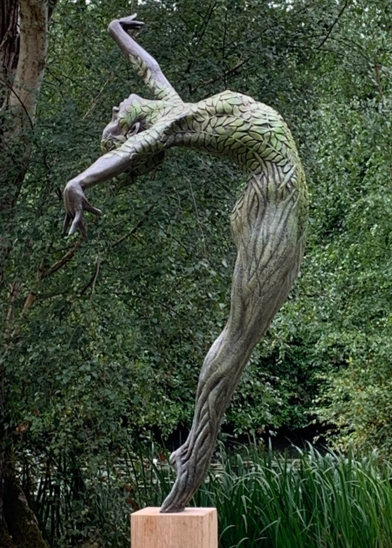 Bronze sculpture of a slender female figure arched forward in a powerful, elongated pose, balanced on one foot atop a wooden plinth. Her body is patterned with leaf-like, cellular shapes that flow from head to toe, echoing natural growth and organic energy. Photographed outdoors among lush green ferns and tall woodland trees.