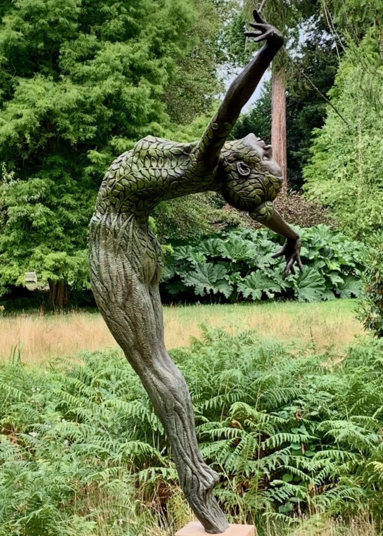 Bronze sculpture of a slender female figure arched forward in a powerful, elongated pose, balanced on one foot atop a wooden plinth. Her body is patterned with leaf-like, cellular shapes that flow from head to toe, echoing natural growth and organic energy. Photographed outdoors among lush green ferns and tall woodland trees.