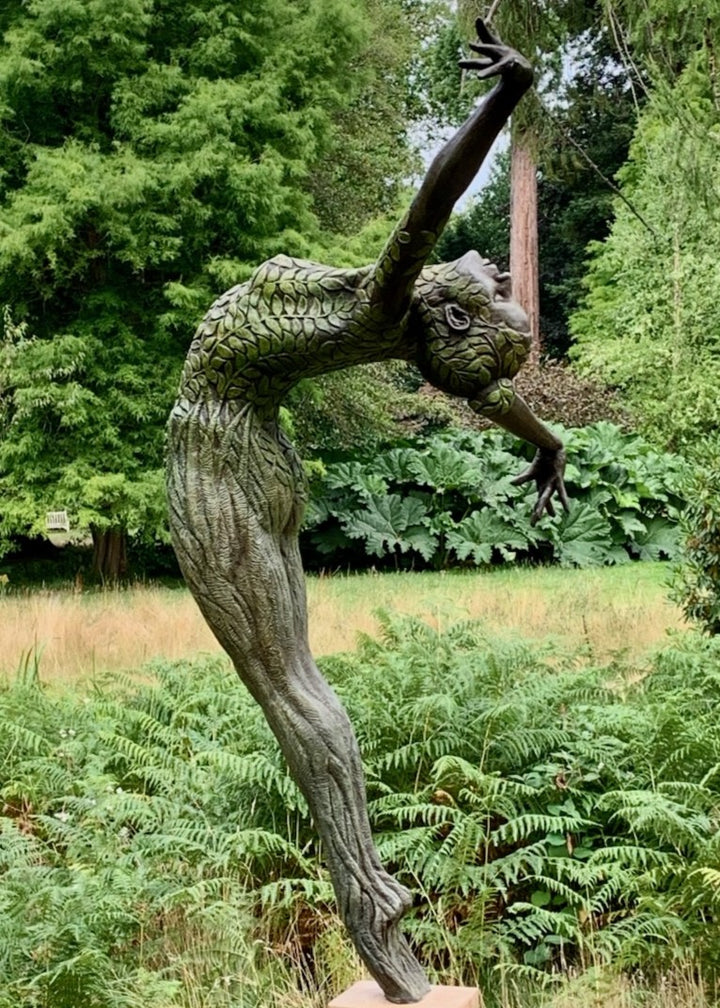 Bronze sculpture of a slender female figure arched forward in a powerful, elongated pose, balanced on one foot atop a wooden plinth. Her body is patterned with leaf-like, cellular shapes that flow from head to toe, echoing natural growth and organic energy. Photographed outdoors among lush green ferns and tall woodland trees.