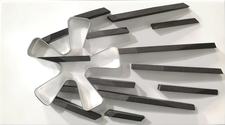 Abstract automotive wall sculpture by Jonny Ambrose featuring a polished aluminium Fuchs wheel with black wooden shards radiating outward to suggest power-slide motion and speed, set on a clean white Corian background with black frame.