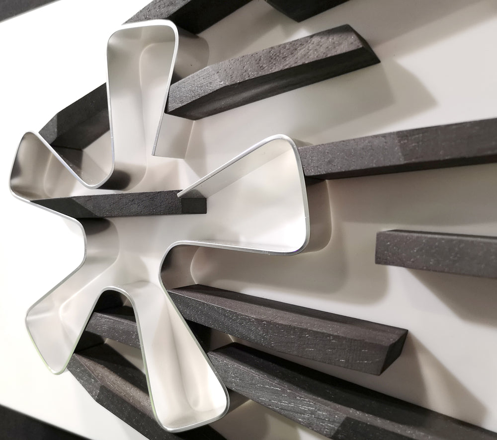 Abstract automotive wall sculpture by Jonny Ambrose featuring a polished aluminium Fuchs wheel with black wooden shards radiating outward to suggest power-slide motion and speed, set on a clean white Corian background with black frame.