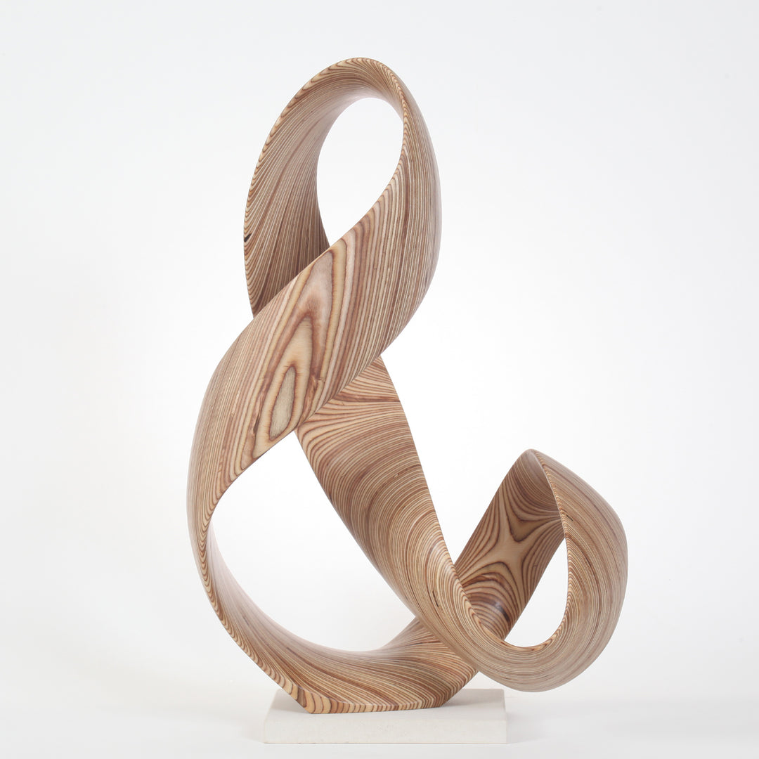 Hand-carved birch plywood sculpture by Bill Prickett, formed as a flowing Möbius-style infinity loop inspired by an ampersand, with continuous twisting curves, visible layered grain, and a balanced sculptural presence on a white base.