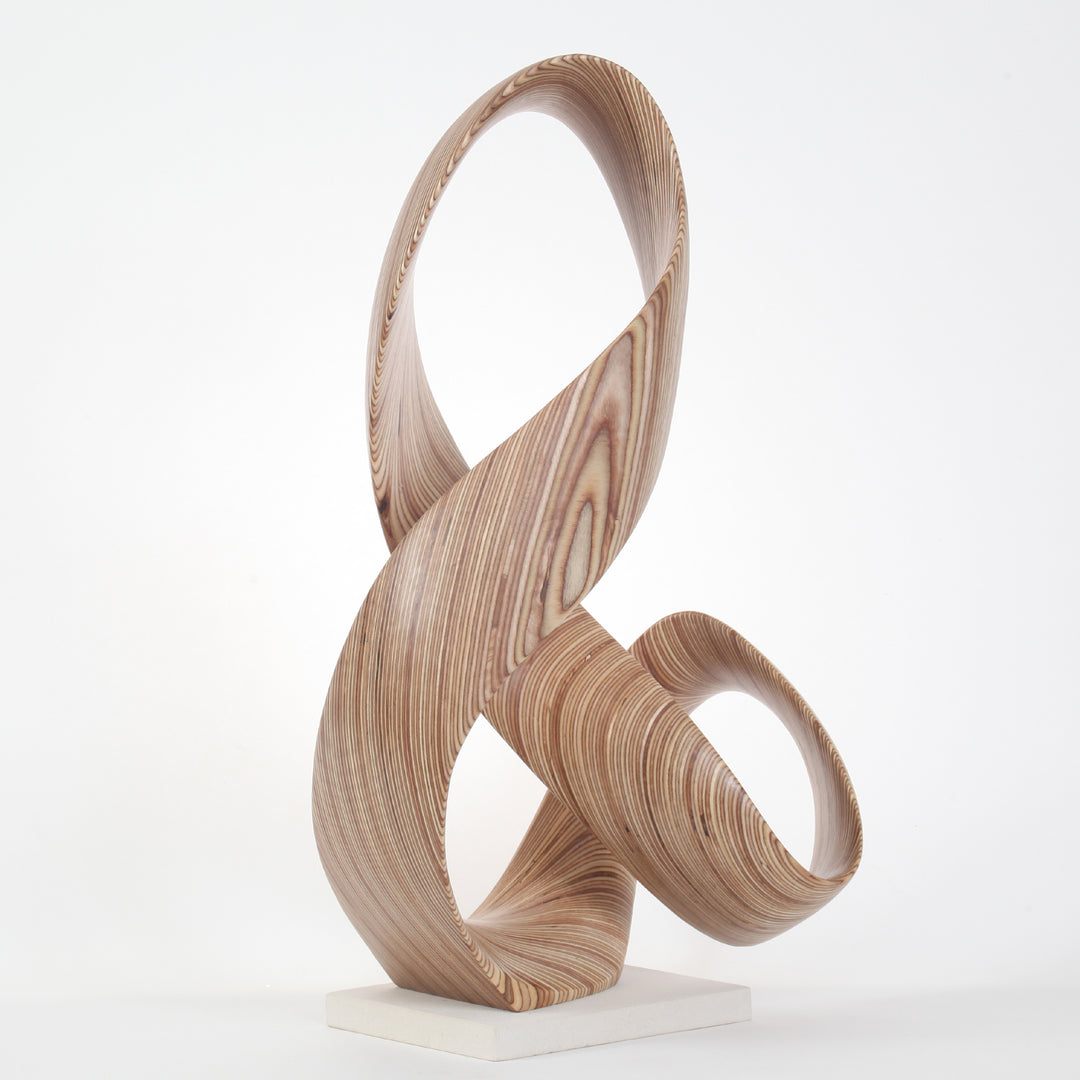 Hand-carved birch plywood sculpture by Bill Prickett, formed as a flowing Möbius-style infinity loop inspired by an ampersand, with continuous twisting curves, visible layered grain, and a balanced sculptural presence on a white base.