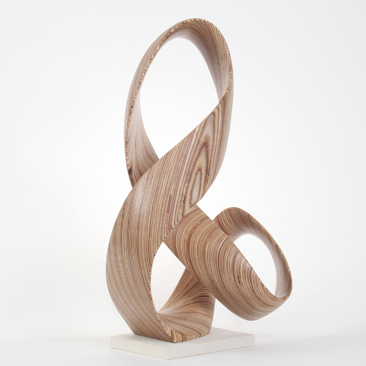 Hand-carved birch plywood sculpture by Bill Prickett, formed as a flowing Möbius-style infinity loop inspired by an ampersand, with continuous twisting curves, visible layered grain, and a balanced sculptural presence on a white base.